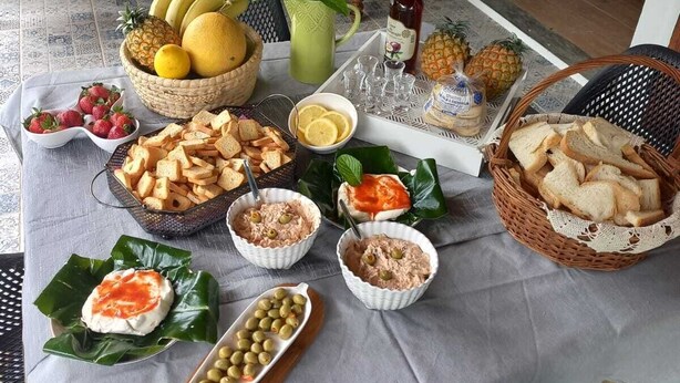 A table set with various foods and drinks, a wicker chair, and a wooden bench in a room with a door and a plant. - Historic Stable House | Private Patio & Farm Views (Lagoa, São Miguel Açores)