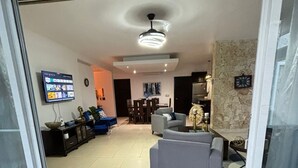 Una sala moderna con un sofá, una mesa de centro y un televisor montado en la pared. Se observa un comedor con mesa y sillas al fondo.