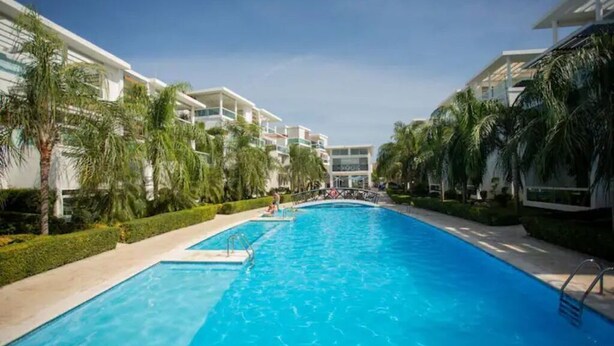 Piscina rodeada de edificios modernos con balcones, palmeras y un cielo despejado.