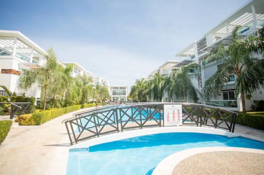 A modern hotel with a central swimming pool, surrounded by palm trees and a bridge.