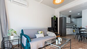 Ein modernes Wohnzimmer mit einem grauen Sofa, einem Glastisch und einem kleinen Beistelltisch mit einer Pflanze. Im Hintergrund ist eine Küche mit weißen Schränken und einem schwarzen Stuhl zu sehen.