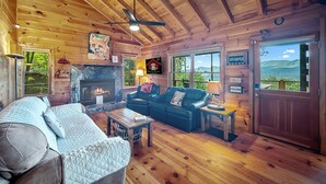 A cozy cabin interior with a fireplace, wooden walls, a ceiling fan, and a view of the outdoors.
