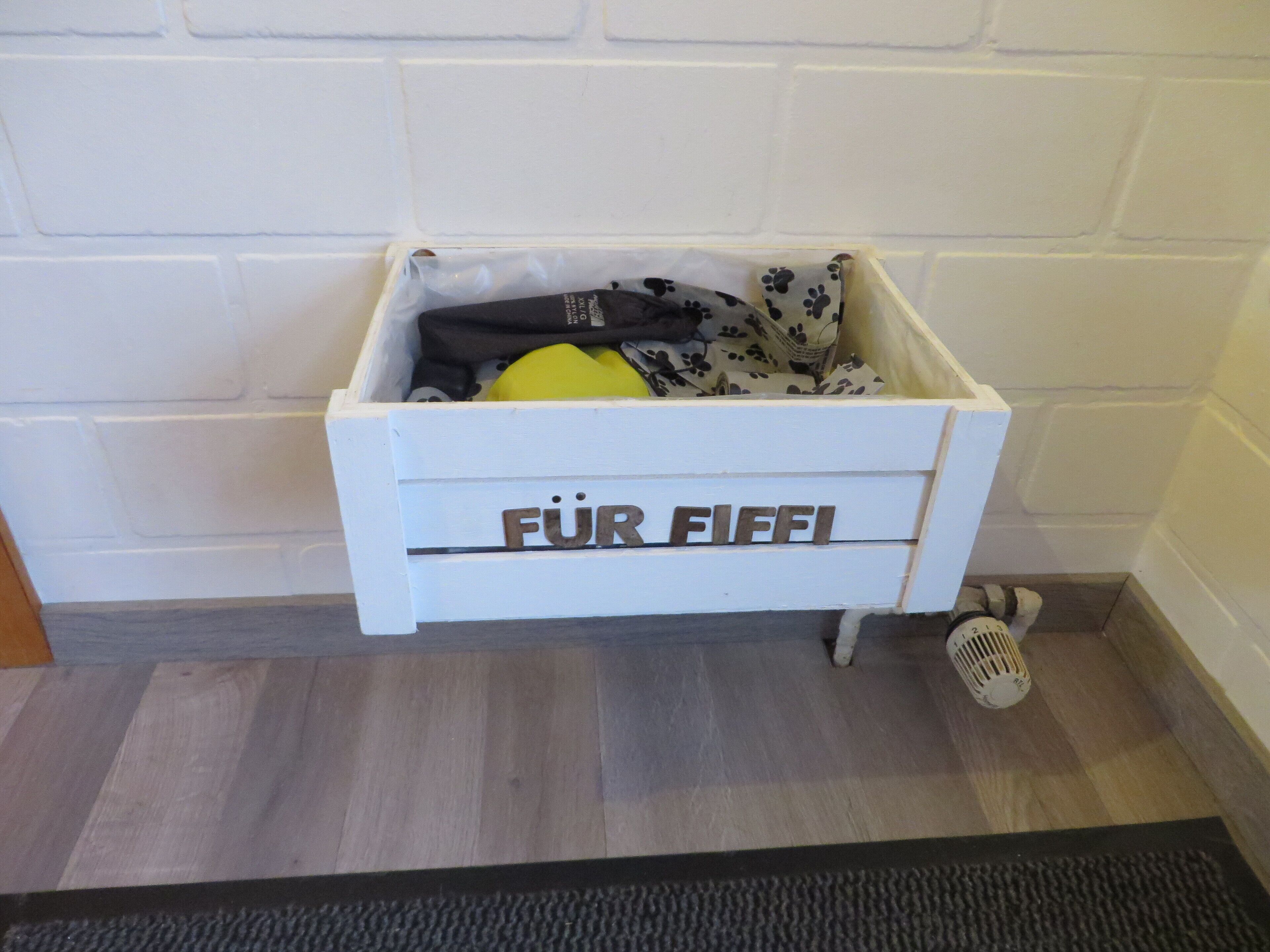 Valkoinen säilytyslaatikko, jonka kyljessä lukee ”FÜR FIEFI”, sisältää keltaisen esineen ja kuviollisen kankaan, ja se on sijoitettu valkoista tiiliseinää vasten.
