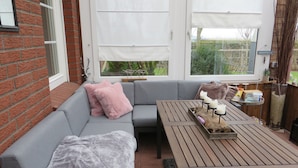 A cozy outdoor seating area with a grey sofa, a wooden table, and a view of greenery through large windows. - Huus Inge and Manni (Dornum)