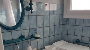 Kylpyhuone, jossa on valkoinen pesuallas, peili, wc ja hylly, jossa on hygieniatuotteita.