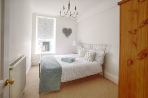 A bedroom with a bed, a radiator, a window with a floral curtain, a chandelier, and a wooden door.