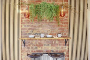 A brick wall with a hanging green plant, a shelf with white dishes, and two hanging light bulbs.
