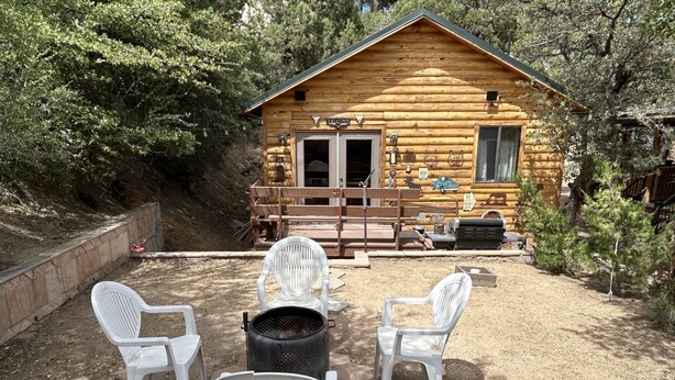 Ein rustikales Blockhaus mit Veranda, umgeben von Bäumen. Vier weiße Plastikstühle sind kreisförmig um einen schwarzen Grill angeordnet.