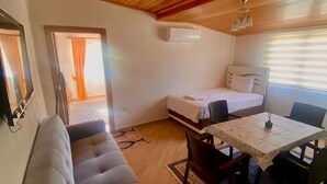 Una habitación compacta con techo de madera, un sofá, cama pequeña, mesa con sillas y alfombra estampada.