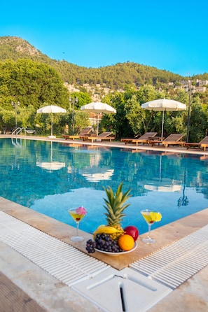 Una piscina con sillas de descanso y sombrillas, un frutero y una bebida en el borde.