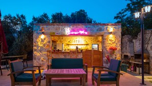 Un bar al aire libre con paredes de piedra, mobiliario de madera y un letrero de neón.