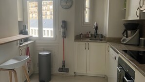 Una cucina compatta con mobili bianchi, una finestra vista edifici, una sveglia, una macchina da caffè e un cestino.
