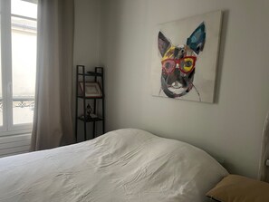 A bedroom with a bed, a wall-mounted painting of a dog, a bedside table with a lamp, and a window with curtains.