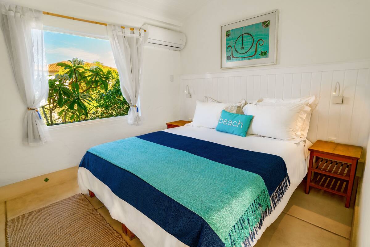 Une chambre à coucher avec un lit, une fenêtre avec des rideaux, un climatiseur fixé au mur, une table de chevet et une œuvre d’art décorative accrochée au mur.