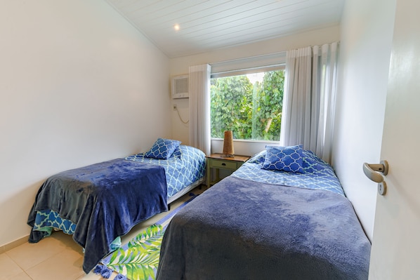 A bedroom with two beds, a window with curtains, a small table with a lamp, and a wall-mounted air conditioning unit. - Pousada Maré (Búzios)