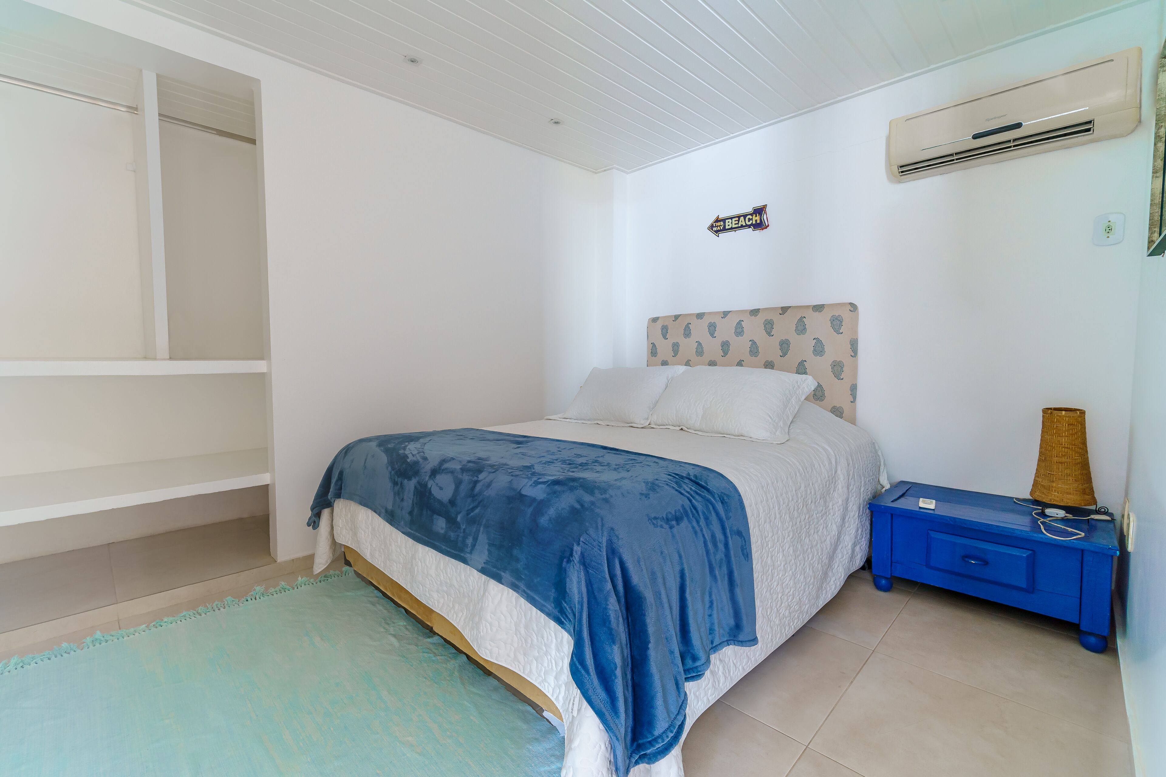 Un dormitorio con cama, mesita de noche, aire acondicionado instalado en la pared y un cartel que indica la dirección a la playa.