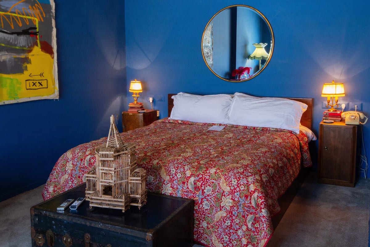 Quarto com uma cama grande, um espelho, duas mesas de cabeceira com luminárias, uma mala e uma gaiola decorativa para pássaros.