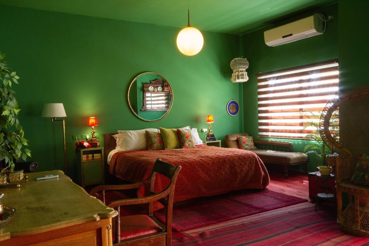 Um quarto com parede verde, um espelho redondo, uma cama com colcha vermelha, uma cadeira de madeira, uma mesa com abajur e uma janela com persianas.