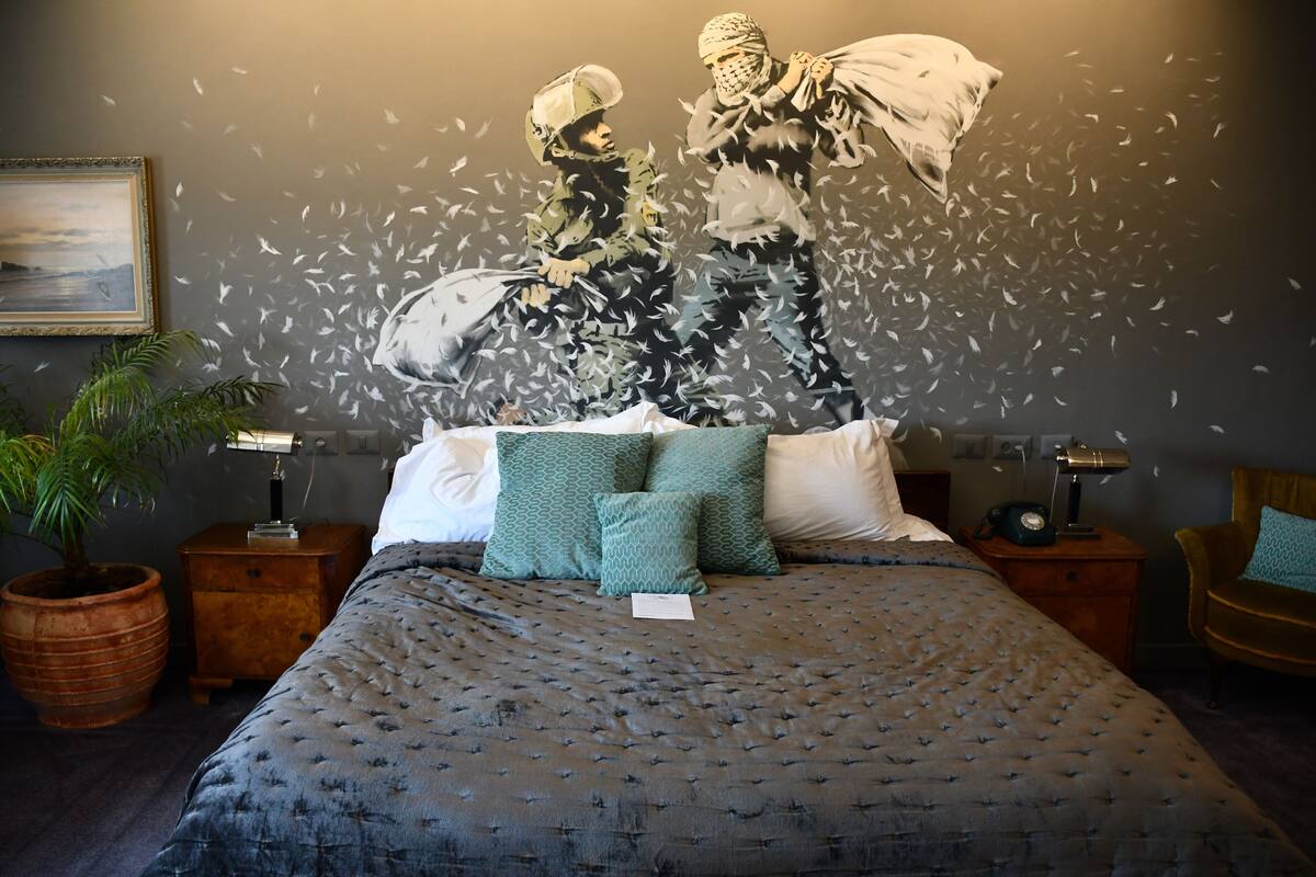 Quarto com cama grande, almofadas estampadas em verde, mesa de cabeceira com abajur, cadeira de madeira e uma planta em vaso grande.
