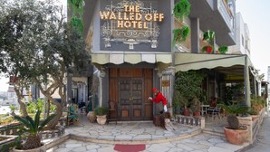 El exterior del Hotel The Wall Off, con un letrero grande, escalones de piedra, plantas en macetas y mesas al aire libre.