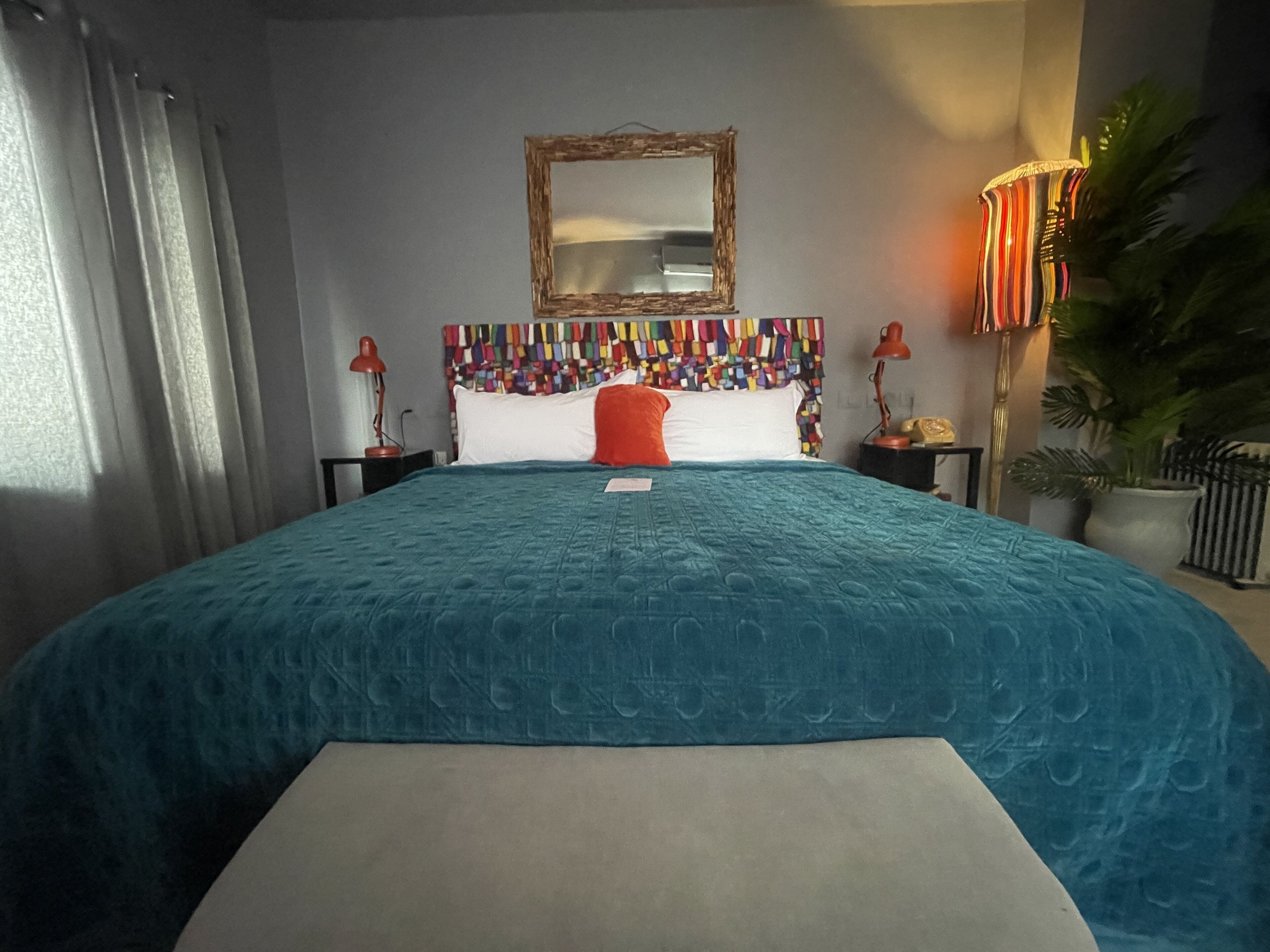 Une chambre d’hôtel avec un grand lit, une tête de lit colorée, un miroir, une table de chevet avec une lampe et une plante en pot.