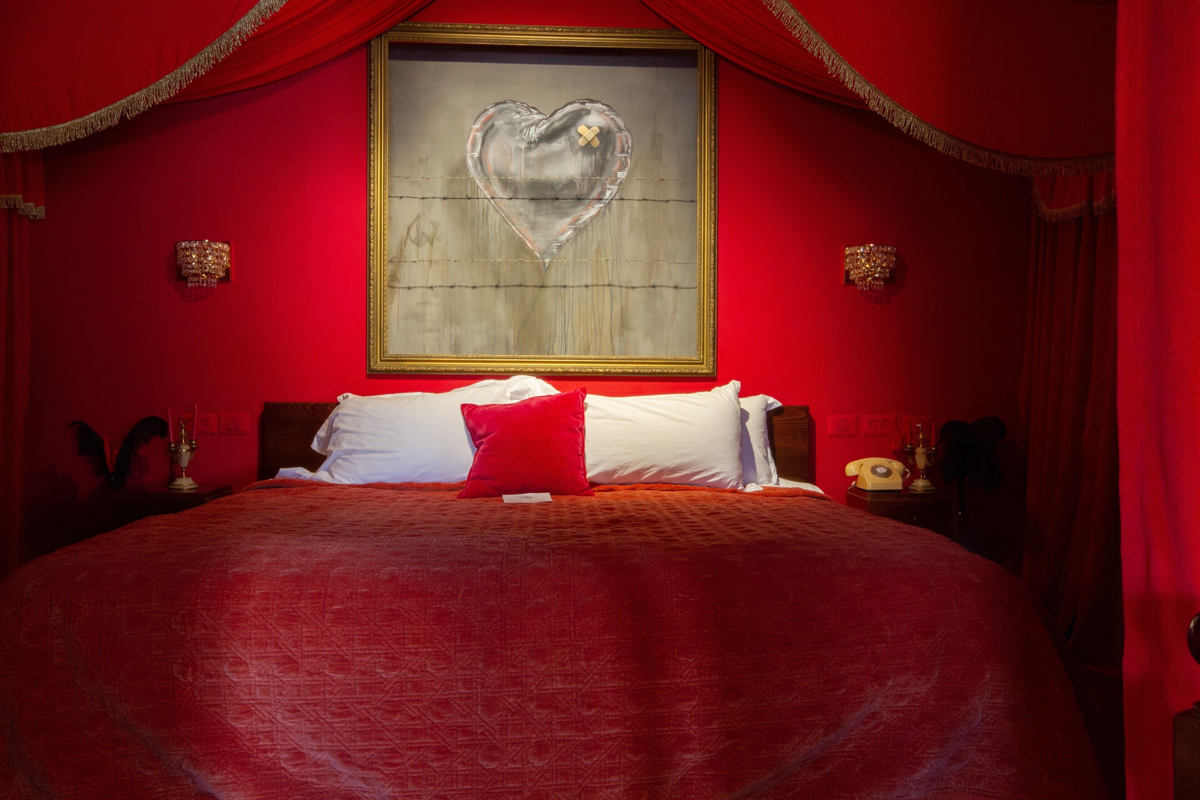A large bed with a red bedspread, two white pillows, and a red accent pillow. There is a framed artwork of a heart on the wall above the bed.