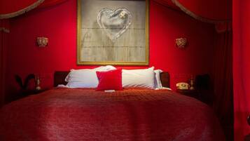 A large bed with a red bedspread, two white pillows, and a red accent pillow. There is a framed artwork of a heart on the wall above the bed.