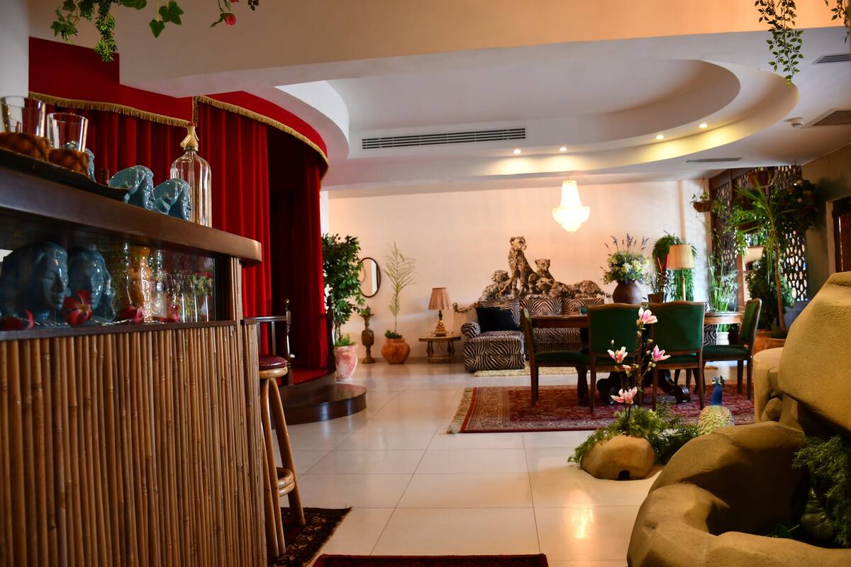 Uma sala de estar espaçosa com um painel de cortina vermelha, um espaço de bar em madeira com objetos decorativos, um sofá estampado com padrão de zebra e diversas plantas em vasos.