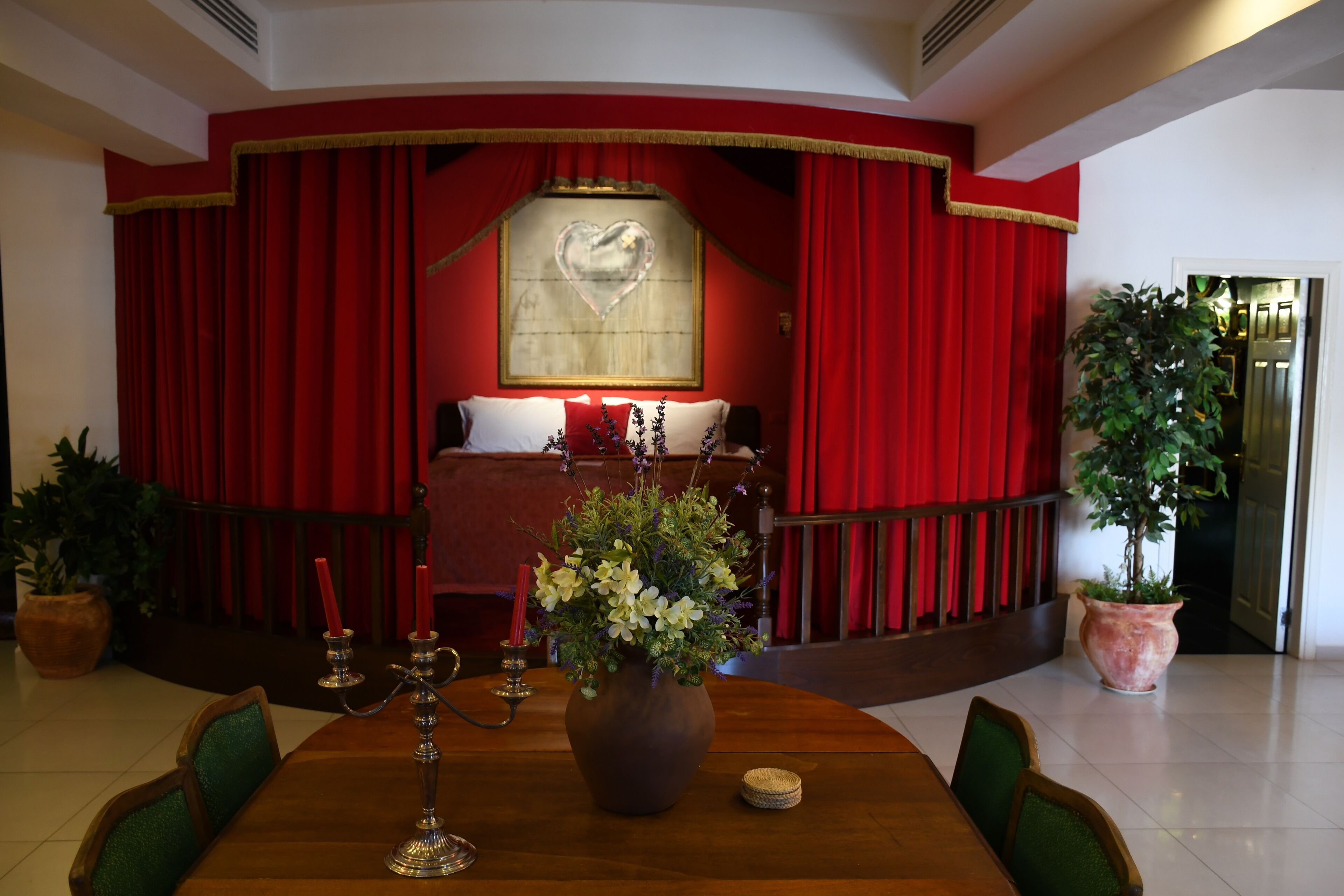 A room with a stage-like area featuring red curtains, a heart-shaped decoration, and a bed with white bedding. There is a wooden table with a vase of flowers and candles, and two potted plants.