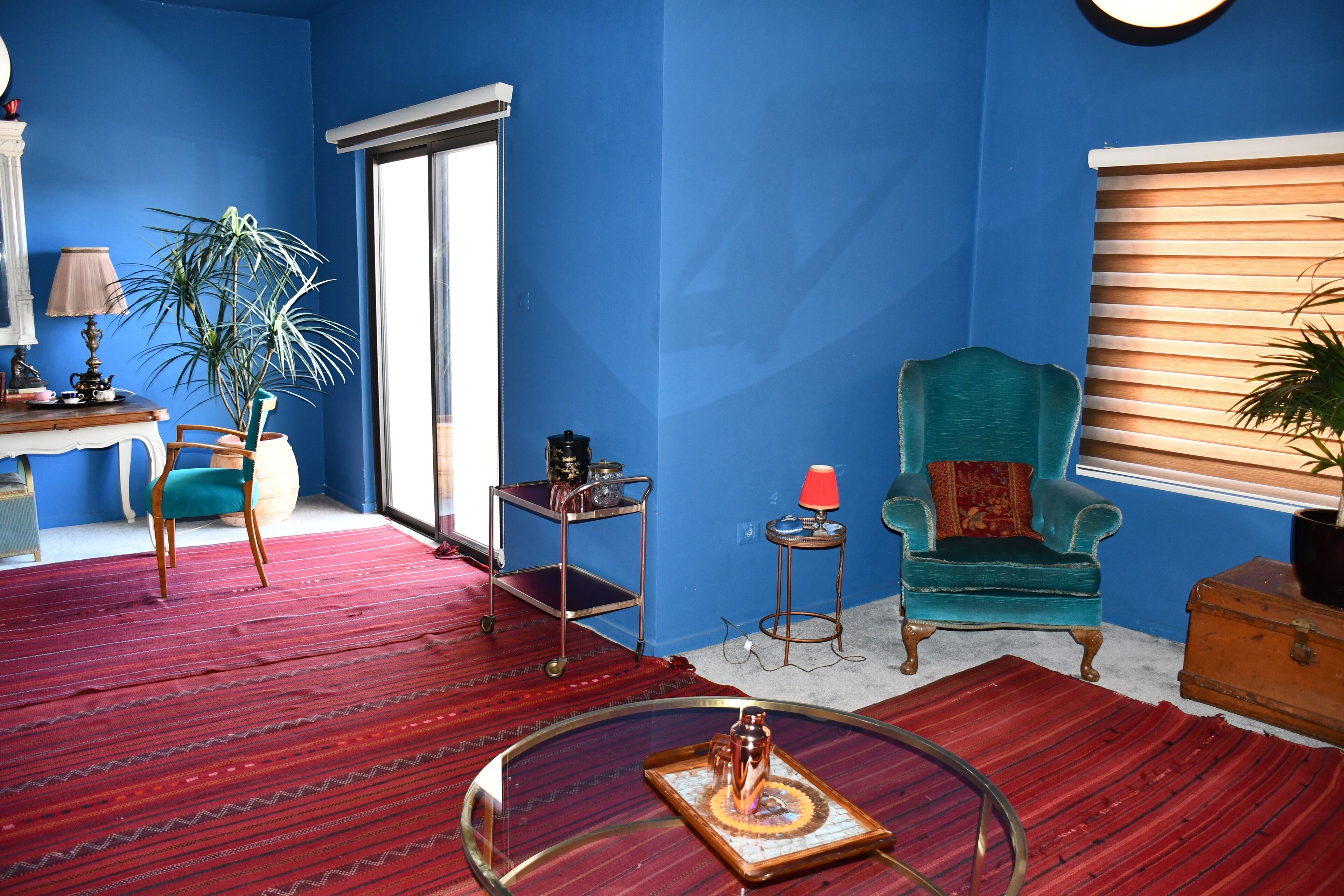 A room with a blue wall, a teal armchair, a red carpet, a glass coffee table, and a wooden cabinet.