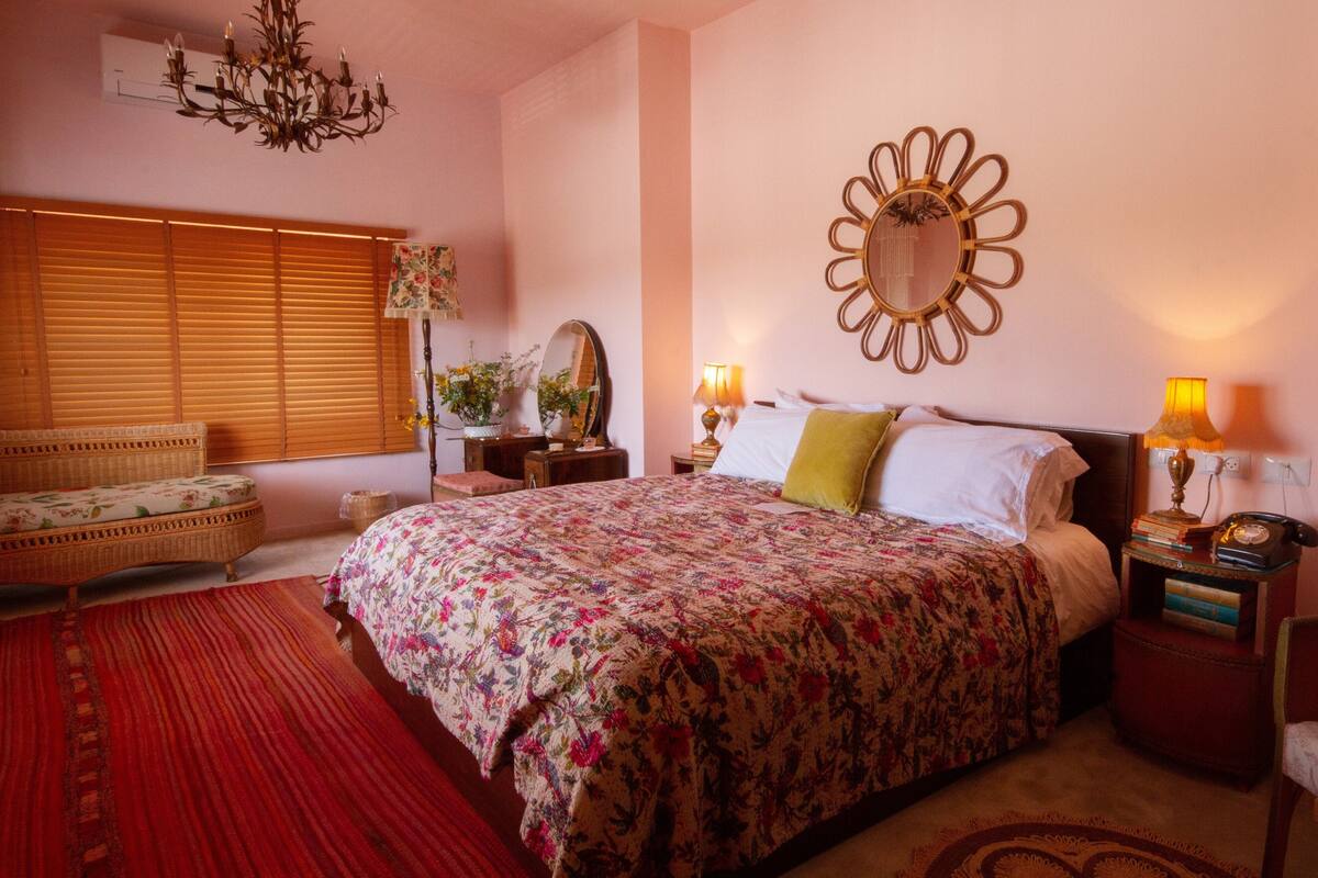 Quarto com uma cama grande, uma mesinha de cabeceira com abajur, um espelho na parede e uma janela com persianas.