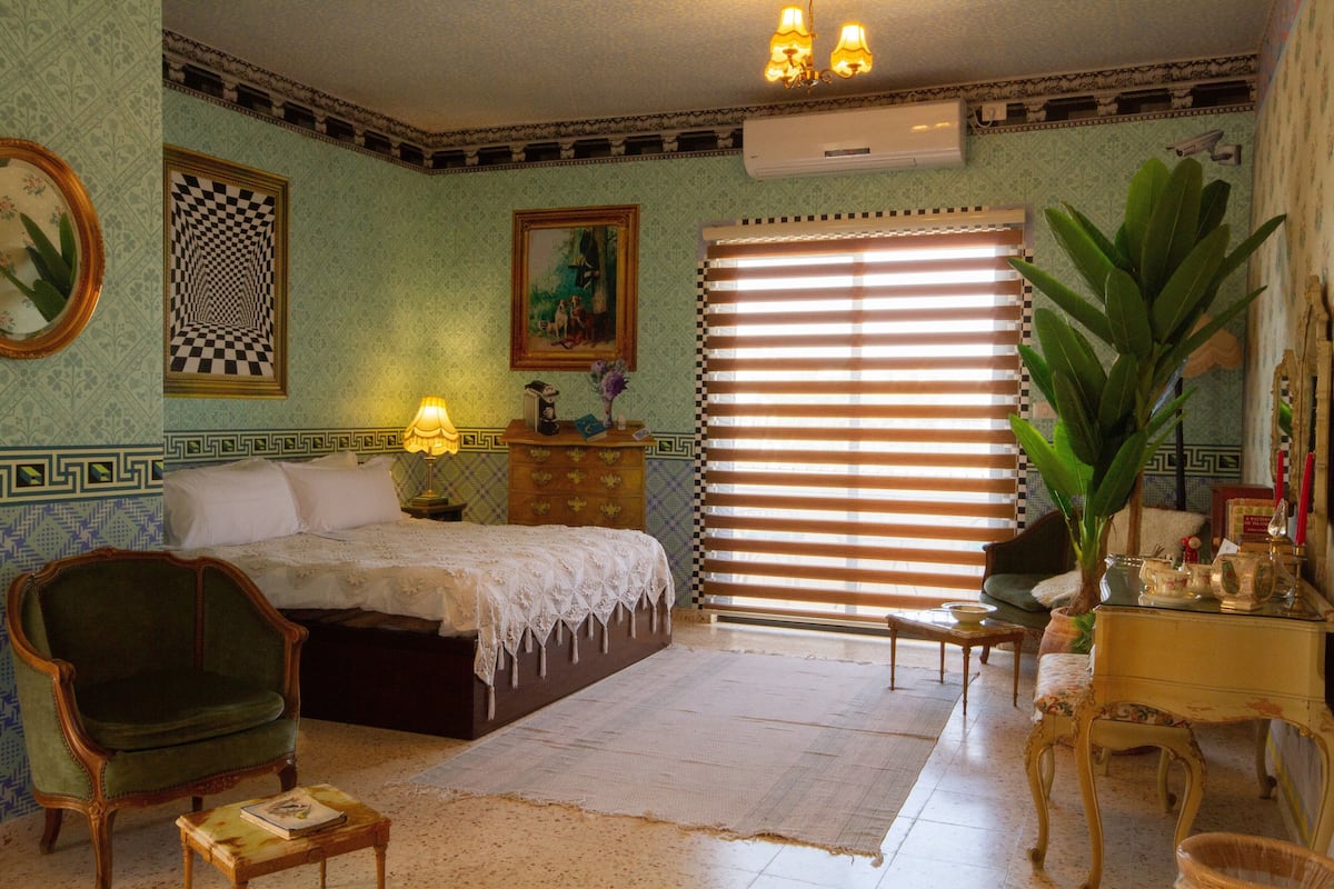 Um quarto com uma cama, uma cadeira, uma mesa, uma luminária e uma janela com persianas.
