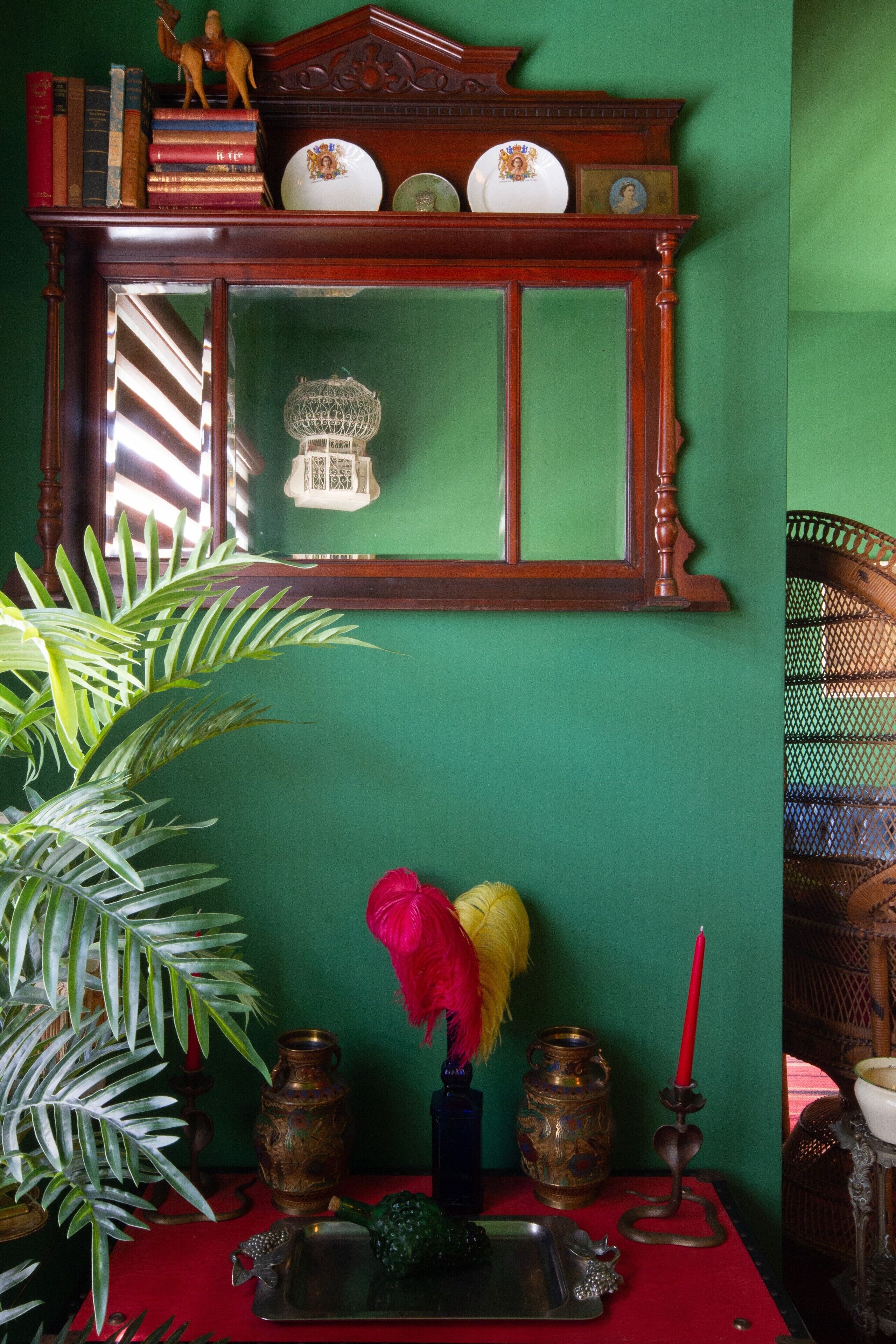 A room with a green wall, a wooden shelf with books and decorative items, a mirror, a red table with a vase, a plant, and a candle holder.