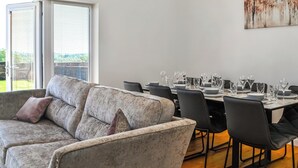 Un coin repas avec une table dressée pour six personnes, un canapé gris et une grande fenêtre avec des stores.