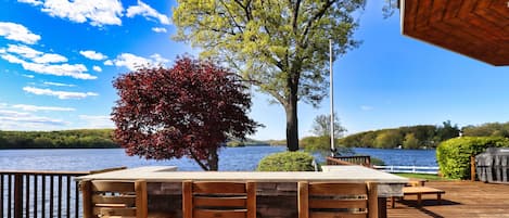 Una terraza de madera con una mesa y sillas de piedra, con vista a un lago con un árbol y un cielo despejado.