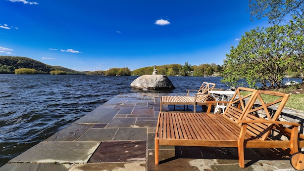 Eine Holzterrasse mit Stühlen und Tisch, mit Blick auf einen See. In der Ferne steht eine Statue auf einem Felsen.