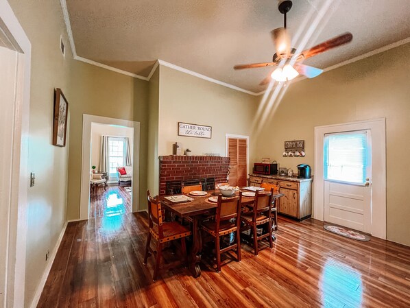 A dining area with a brick wall, wooden table, and chairs. There is a ceiling fan, a kitchen area with a microwave, and a door with a sign.