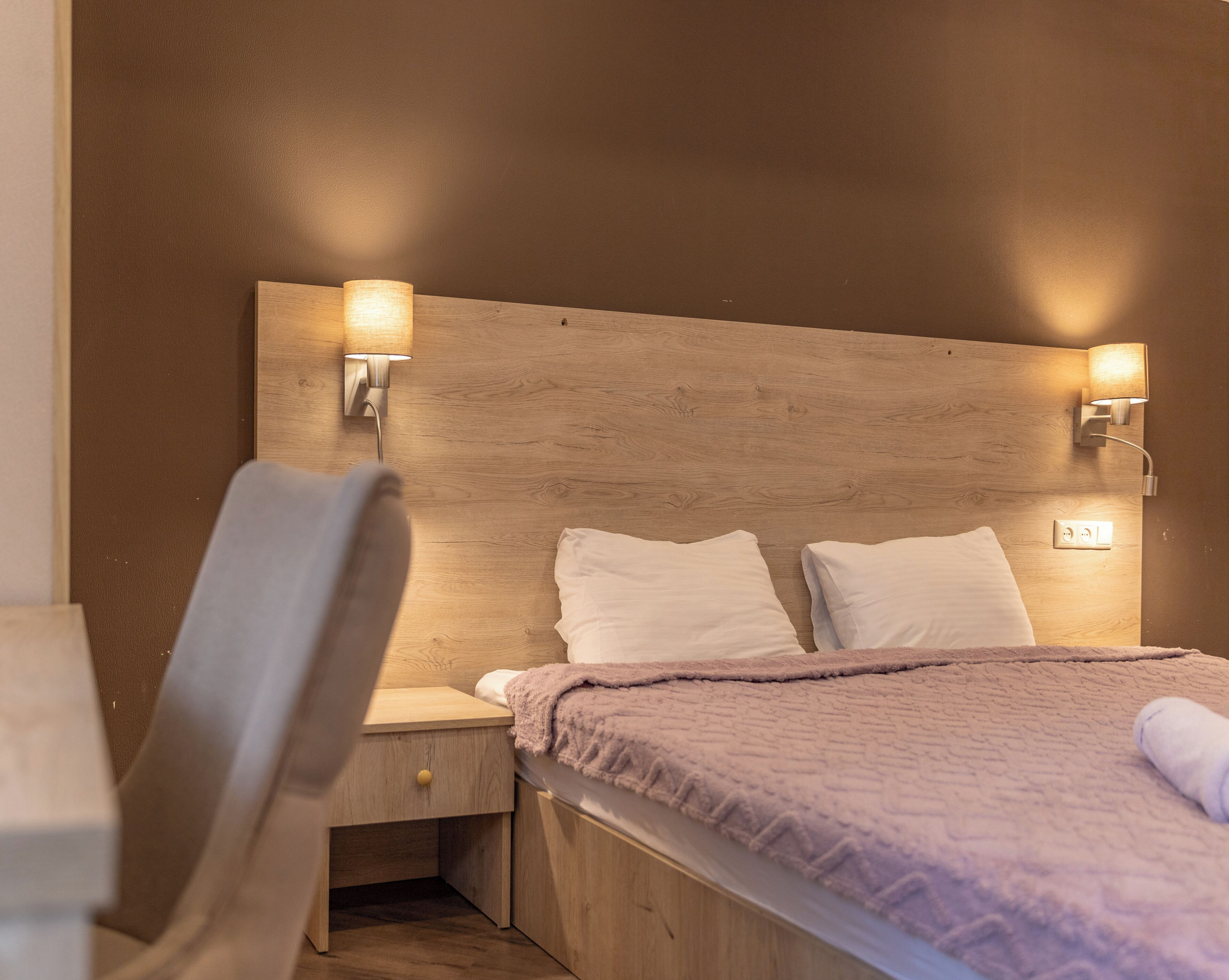 Ein Schlafzimmer mit einem hölzernen Kopfteil, zwei Nachttischlampen, einem Bett mit weißer Bettwäsche und einem Sessel.