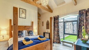 A bedroom with a wooden four-poster bed, a large window with curtains, a small table with a vase, and a view of a stone building outside.