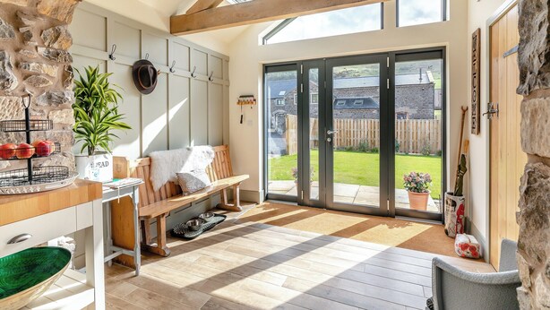 A cozy room with a stone wall, wooden beams, a bench, a dining area with a bowl and a plant, and a view of a garden through a glass door.