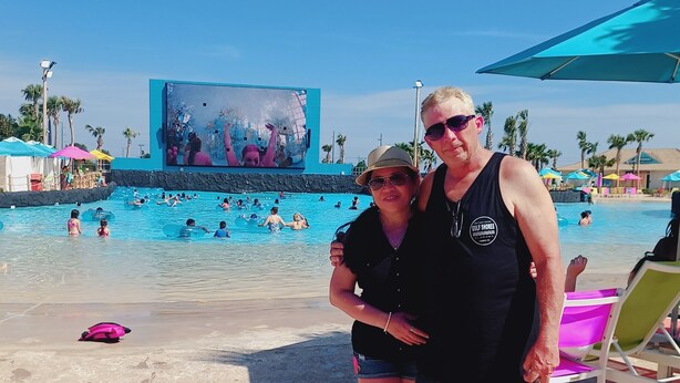 Zona de piscina frente al mar, con una pantalla grande que muestra una multitud, rodeada de palmeras y sombrillas.
