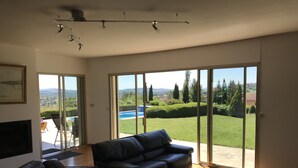 Un vaste salon doté d’un canapé en cuir noir, d’une table basse en verre et d’un tapis rouge. De grandes portes-fenêtres coulissantes donnent sur un jardin avec une piscine et une vue sur la campagne.