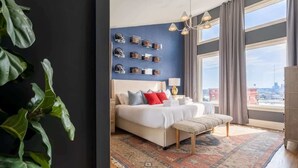 Una moderna camera d'albergo con un letto grande, un comodino, una panca, un'ampia finestra con tende e vista sulla città.