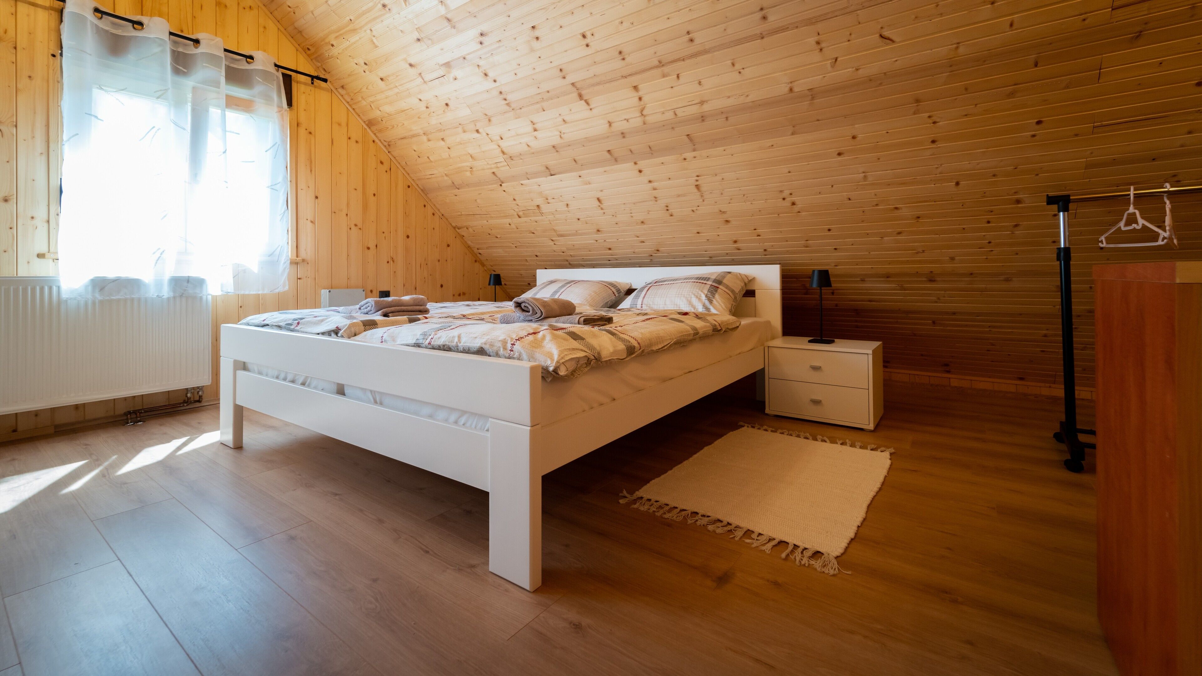 Una camera da letto con soffitto spiovente, pareti in legno, un letto bianco, un comodino e un termosifone.