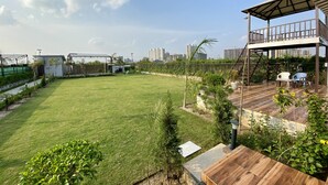A spacious backyard with a wooden deck, a covered seating area, and a view of a cityscape in the background. - GoBravo 71: 2Bhk Farm with Pool & Garden Sector 150 Noida (Noida)