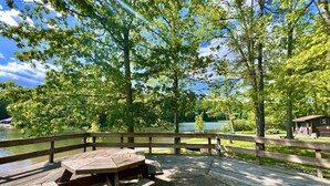 Una terraza de madera con mesas de picnic da a un lago, rodeada de árboles y una valla de madera.