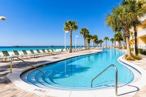 Una piscina con sillones de descanso y palmeras, un muelle que se adentra en el agua, y un cielo azul despejado.