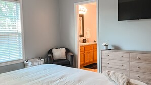 Une chambre à coucher avec un grand lit, une chaise, une commode en bois, un téléviseur et une salle de bain visible à travers une porte ouverte.