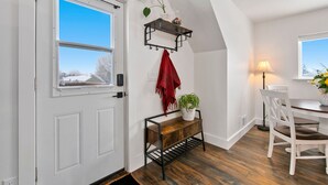 Une pièce avec une porte blanche, un banc en bois orné d’une plante, une serviette rouge, une chaise blanche et une table avec une lampe.