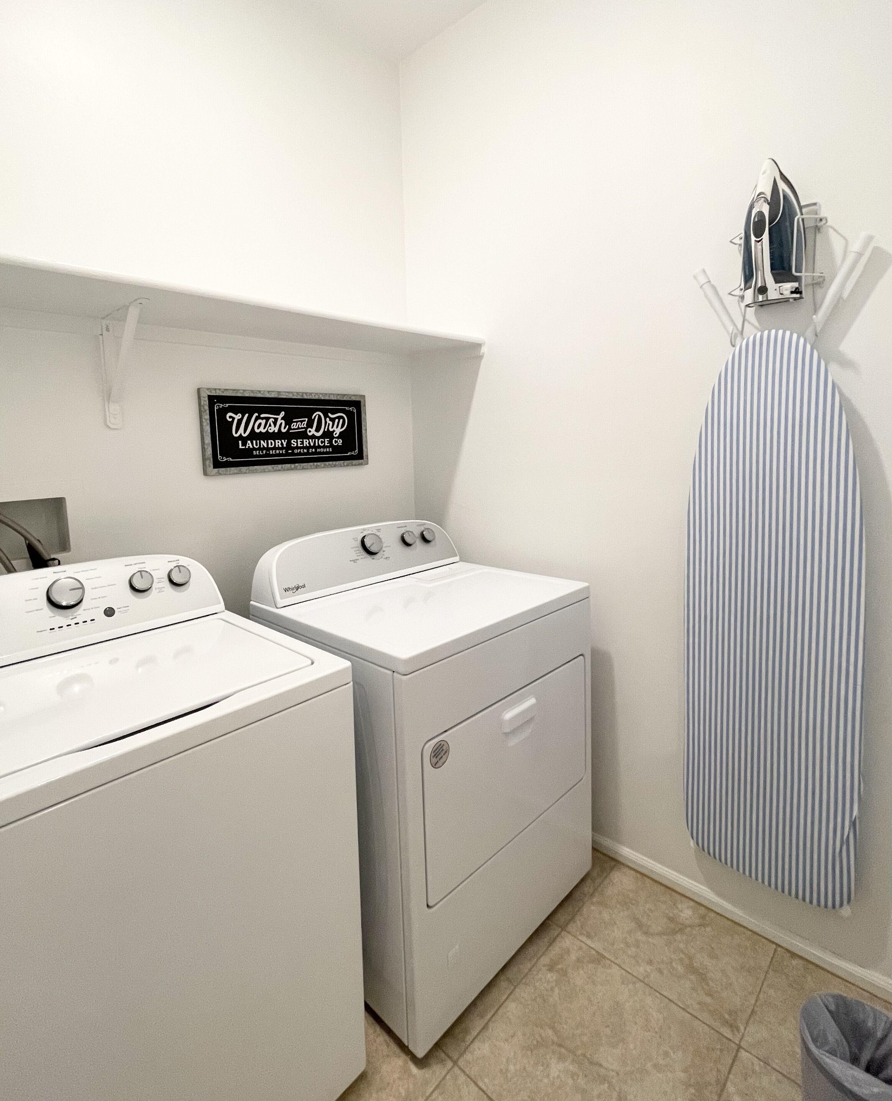 A laundry room with two front-loading washers and dryers, a striped ironing board, and a sign indicating the room is self-serve and open 24 hours.